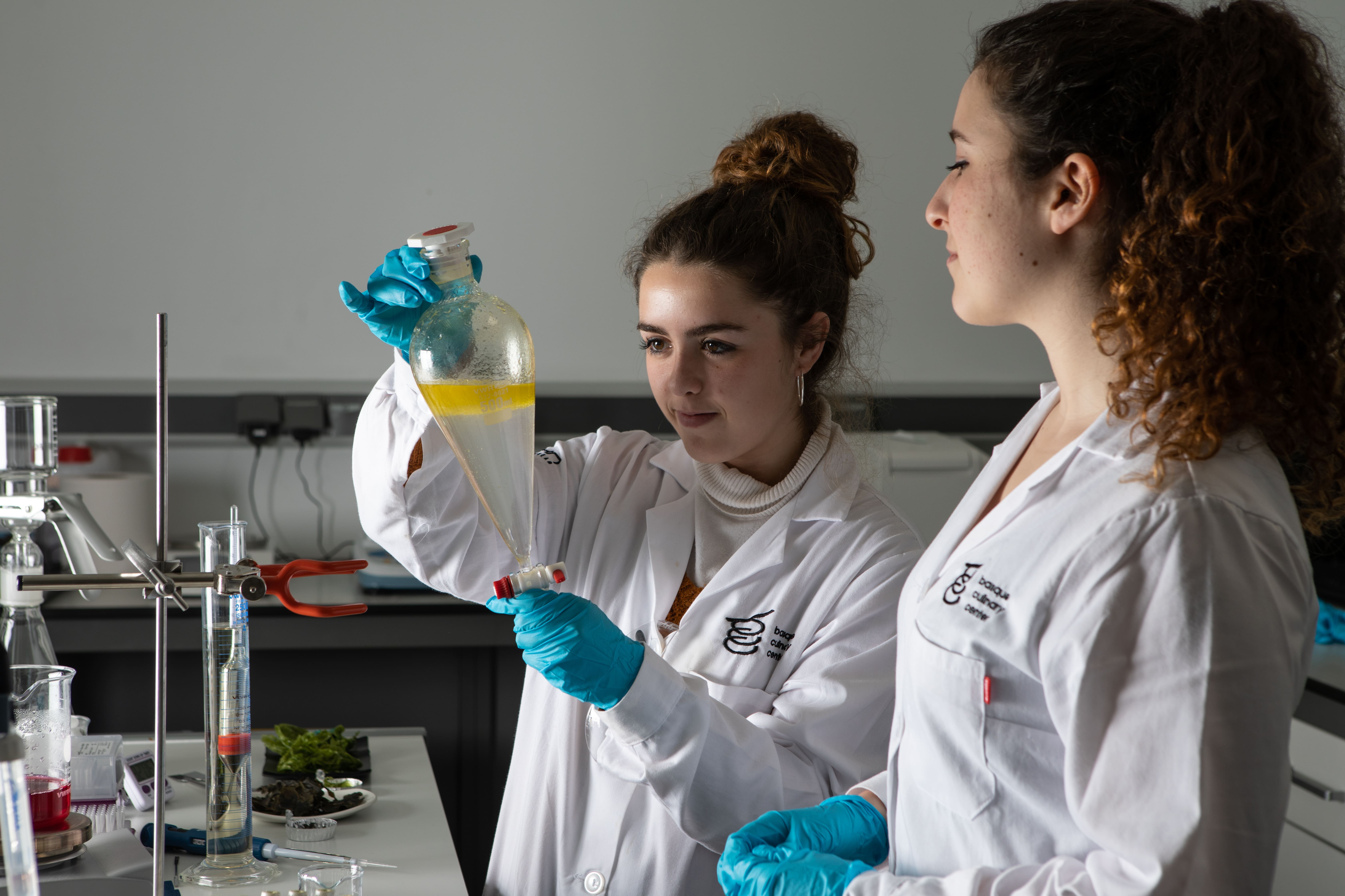 Dos mujeres jóvenes con bata blanca en un laboratorio en la clase del máster de Ciencias Gastronómicas